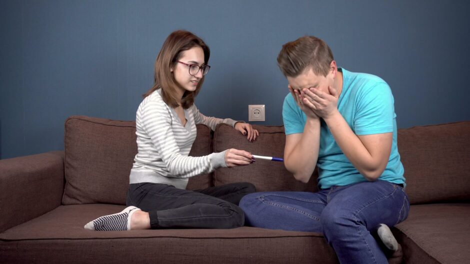 Woman holding pregnancy test, man looking devastated with hands covering his face as he learns it was fake