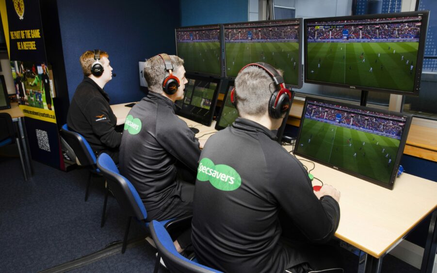 Three referees wearing headphones sit in front of screens showing the football pitch.