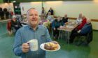Tony Grogan standing with a cup of tea and plate of treats with members of Cuppa Club Aberdeen in the background