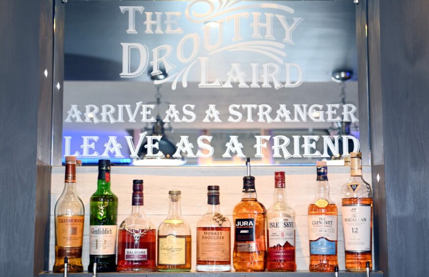 A shelf with bottles of whisky in The Droughty Laird hangs under a mirror that reads "The Drouthy Laird, arrive as a stranger leave as a friend".