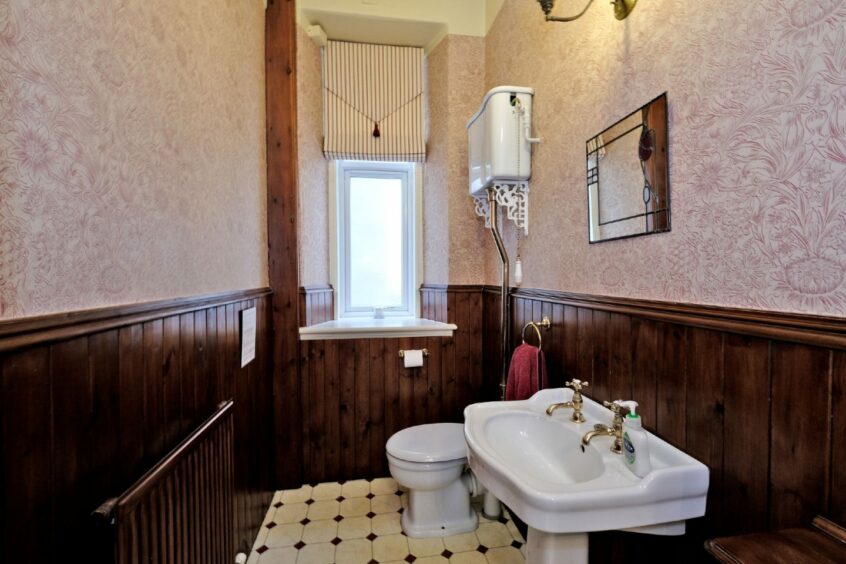 The bathroom with pink elaborate wall paper, dark wood features and a window.