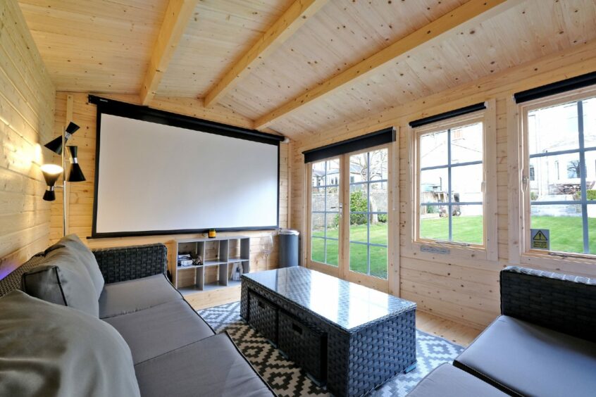 The garden room is made of light wood and it has a projector screen, two outdoor sofas and matching table.