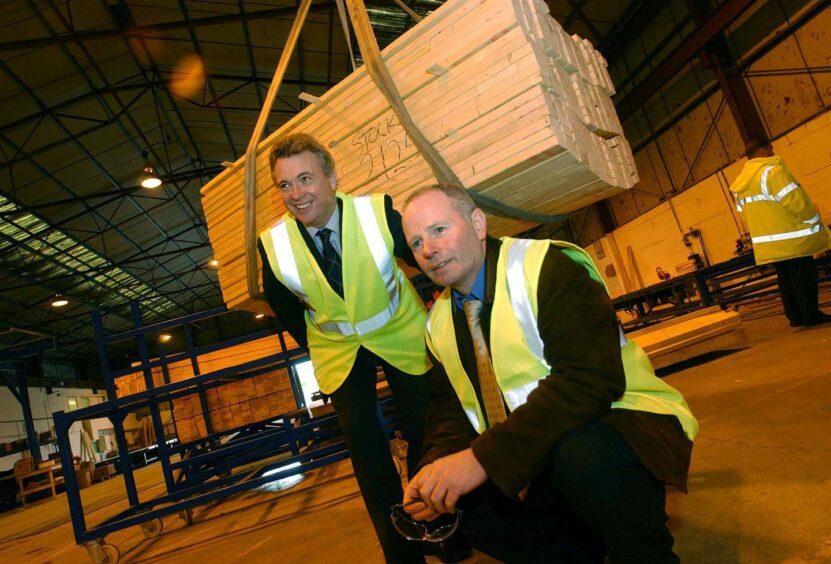 Stewart and Glenn, in hi-vis vests, crouching in front of a stack of timber being lifted. 