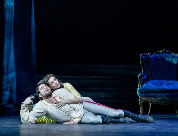 Dancers lying on the stage, with the woman hugging the man from behind. He is holding a gun prop to his head