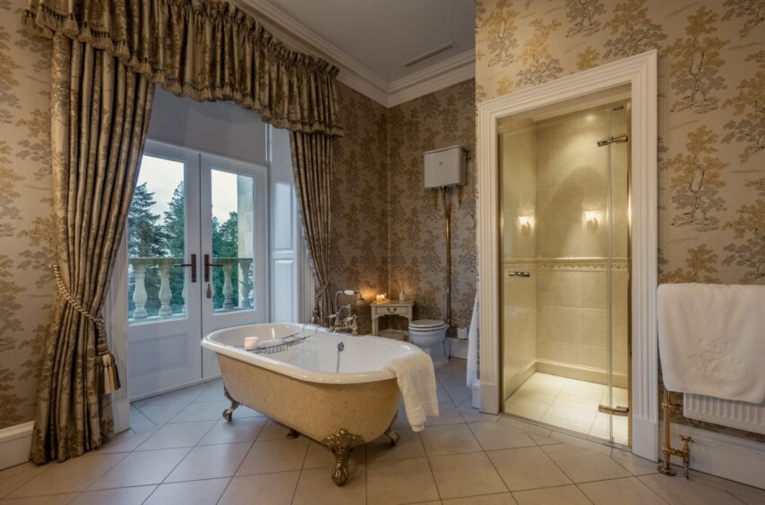 A grand bathroom within Dalhebity House, with a freestanding claw footed tub and encased glass shower. It leads to a balcony.