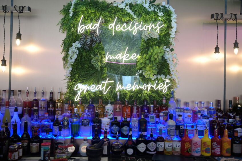 Behind the bar at Jimmy Badgers, a neon sign on greenery reads "Bad decisions make great memories" above shelves of alcohol. 