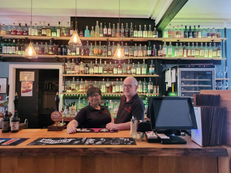 Former owners of Kinlochewe Hotel Karen and David Twist leaning against the bar, with four long shelves lined with alcohol behind them. 