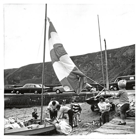 At Stonehaven Harbour a group of people prepare a small boat while a two year old hauls the sail in 1970
