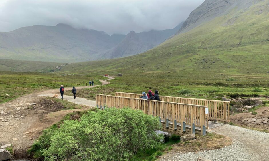 A new path has been built to the Fairy Pools over a bridge rather than stepping stones over the river. The picture shows the glen with people walking on the paths. 