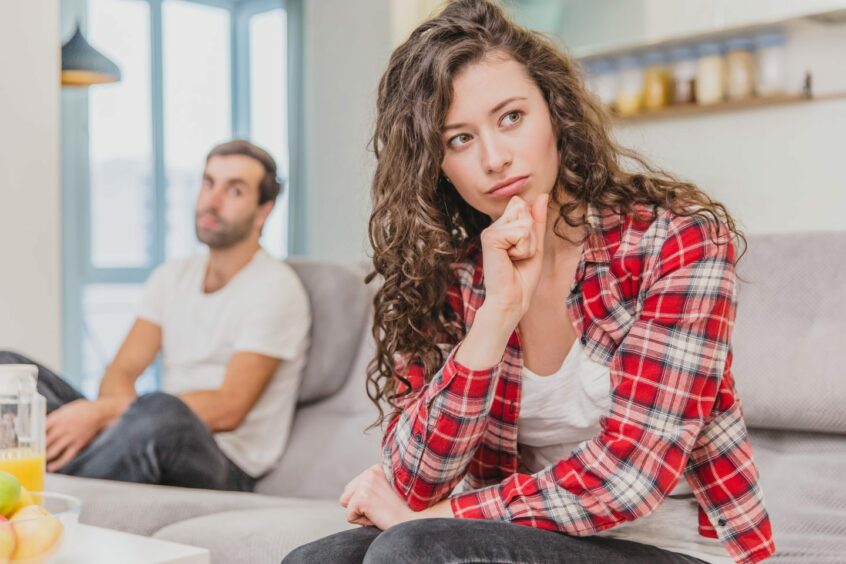 Woman giving man the silent treatment as she turns her back from him while he's talking to her on the sofa