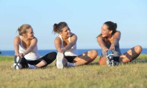 Group of three women stretching after sport on the grass with the sea in the background