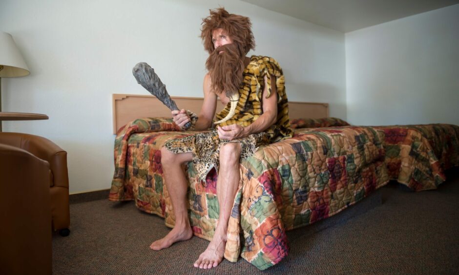 Bored caveman sitting indoors on the edge of a bed with a colorful bedspread in an anonymous cheap motel room