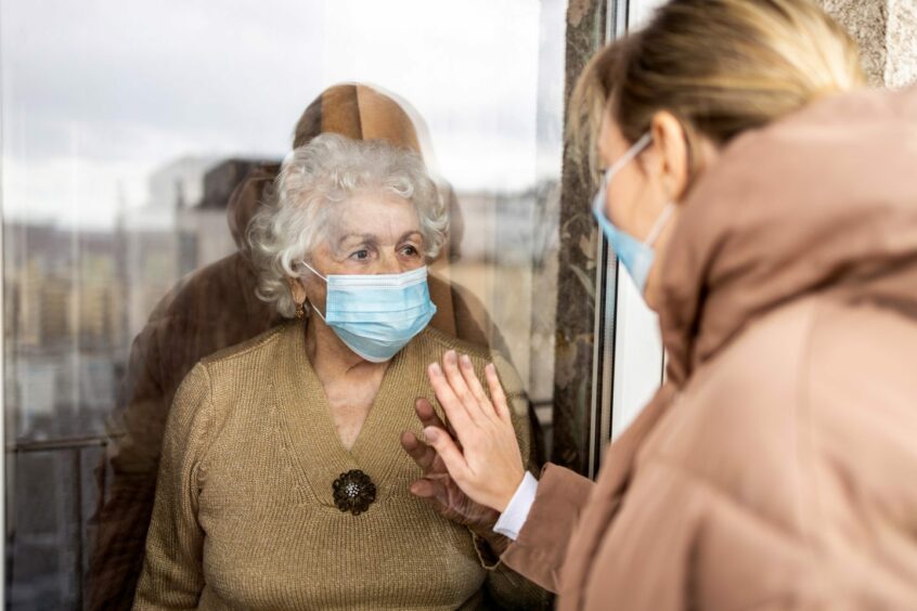 old woman wearing facemask looking out window at daughter also in a mask during lockdown