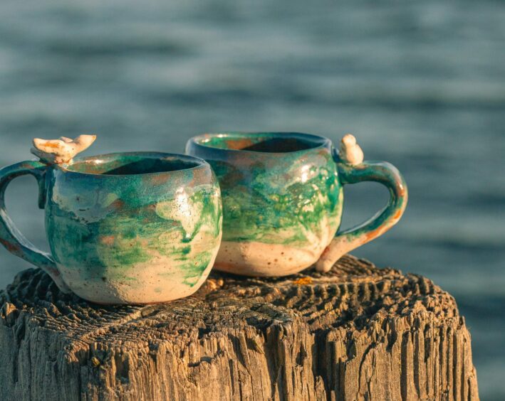 Two handmade ceramic mugs on a wooden post with sea in the background