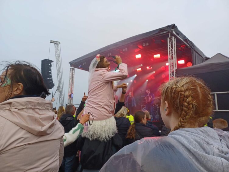A bride-to-be in a hen party outfit rides on her friend's shoulders in front of a festival stage.