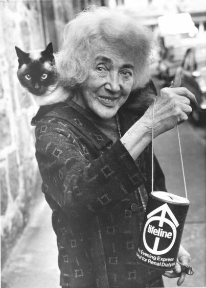 Sunbeam, a Siamese cat, lying across Meg Mann's shoulders outside, while she lifts a donation tin. 