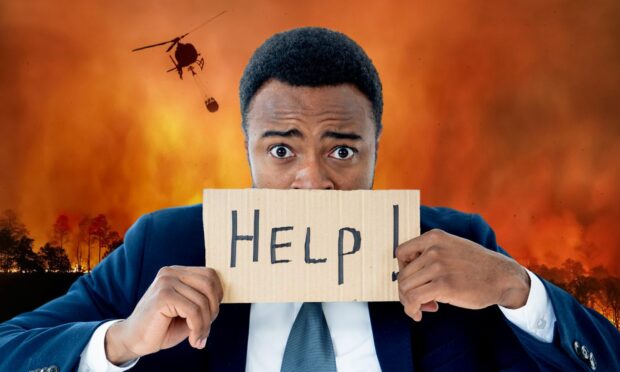 Man holding up cardboard sign with 'help' written on it and behind him is a wildfire with a helicopter coming to spray water on burning forest