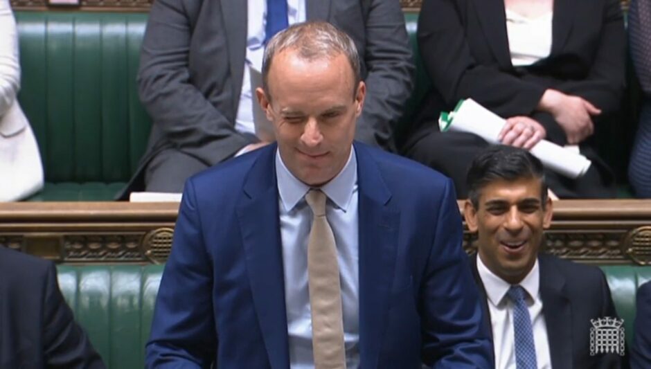 Dominic Raab standing and winking in the House of Commons, as Rishi Sunak smiles sitting down.
