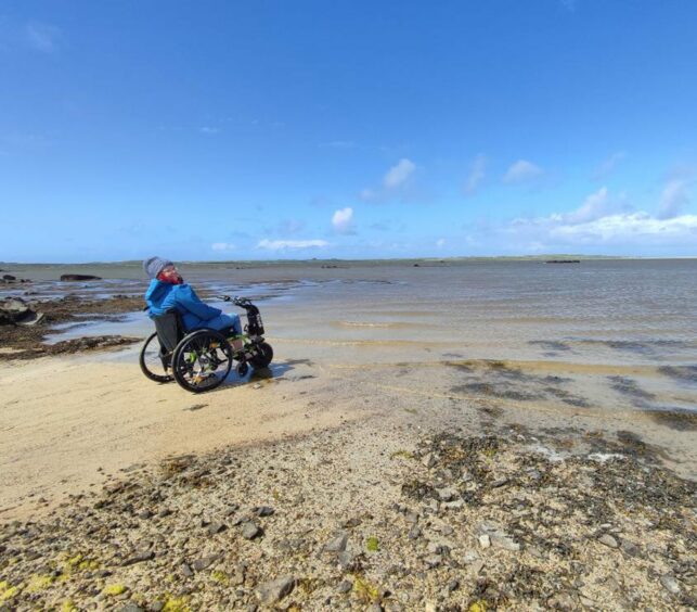 A woman in a wheelchair looks out over a sandy beach,
