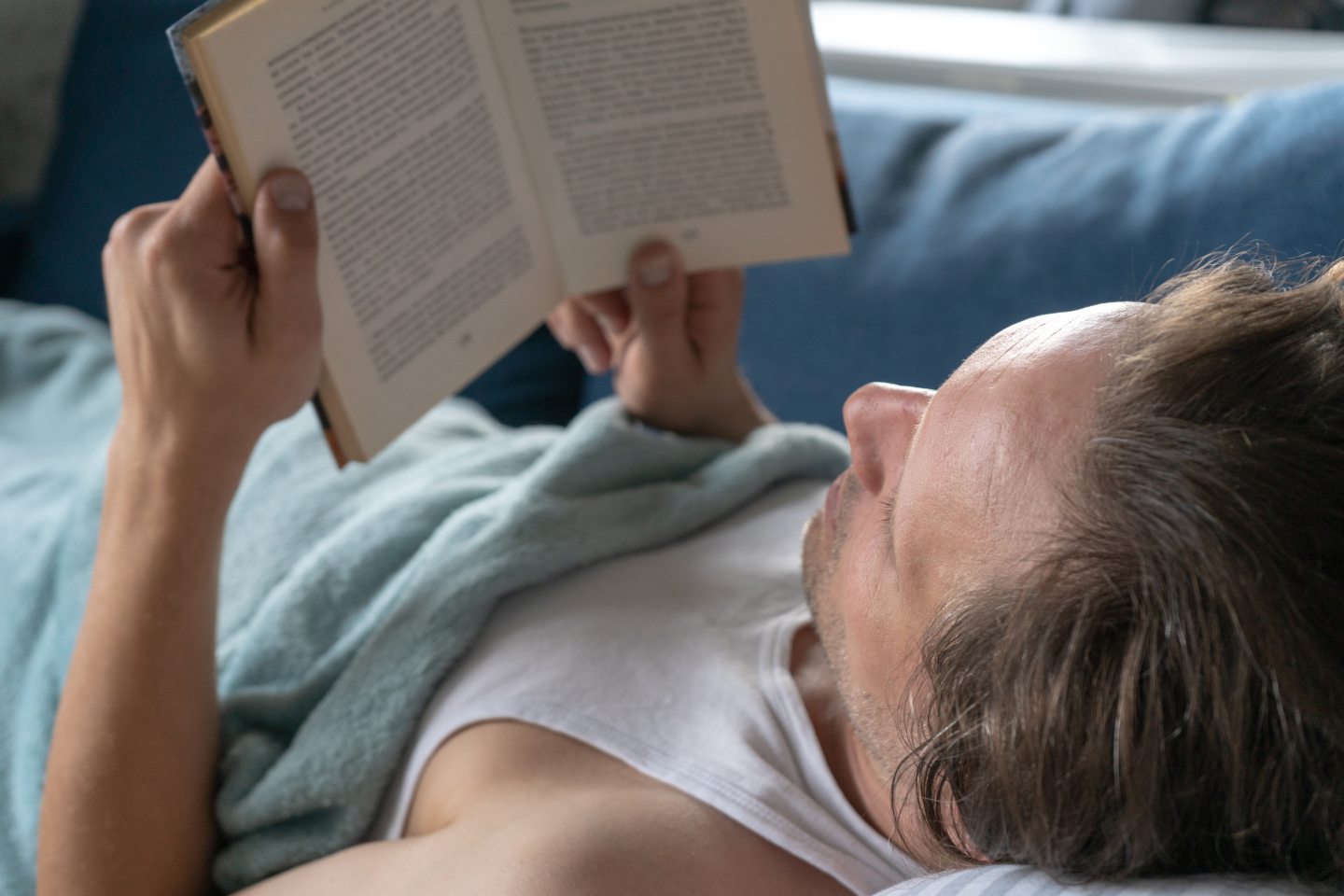 Man lying on the couch with blanket over him reading a book. One of Anne's tips for getting to sleep with insomnia is to wind down in the evening with a laid-back activity like reading.