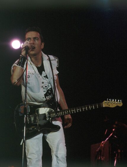 Joe Strummer leaving forward in to the microphone, with his guitar at his waist. 