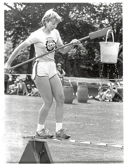 A woman stepping across a seesaw obstacle with a bucket of water on a stick in her hands during a contest at It's a Knockout