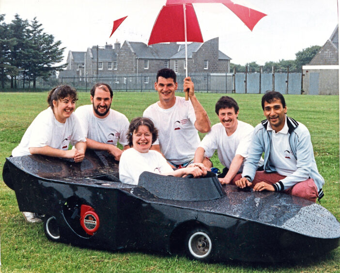 A team surrounding a black go kart in the rain, one member is in the kart and another is holding a large umbrella at It's a Knockout
