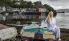 Helen Fields sat on an overturned boat on the shores of Mull, with colourful houses in the background.