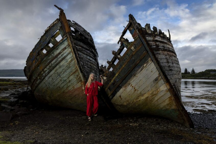 Helen Fields, in a bright red outfit, leaning against the wrecks of two beached fishing boats. Salen Bay, Mull. 