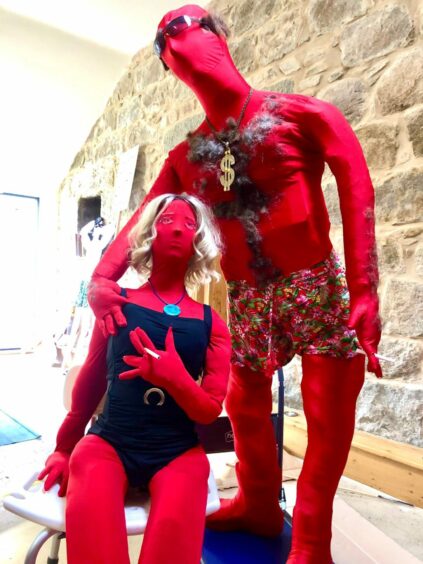 Two stuffed red morphsuits. One sits, decorating with a blonde wig, in a black swimming costume, and blue pendant. Posed standing to the right, the other suit features oversized sunglasses, with hair stuck on its head to mimic male pattern baldness. It also features a hairy chest, with a large dollar sign necklace and floral shorts. They both have cigarettes attached to their hands.