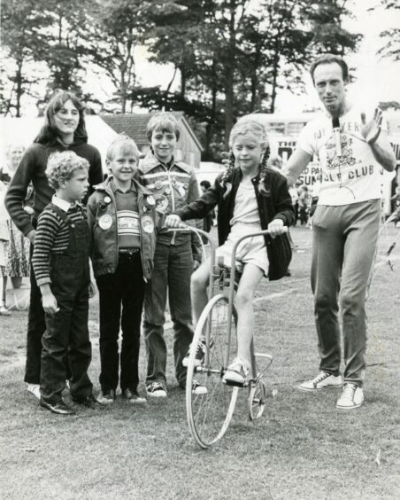 1981- Holiday girl Kimberley Stewart, 6, from Maine, United States, tries out a penny-farthing, watched by comedy unicyclist Roy Rivers