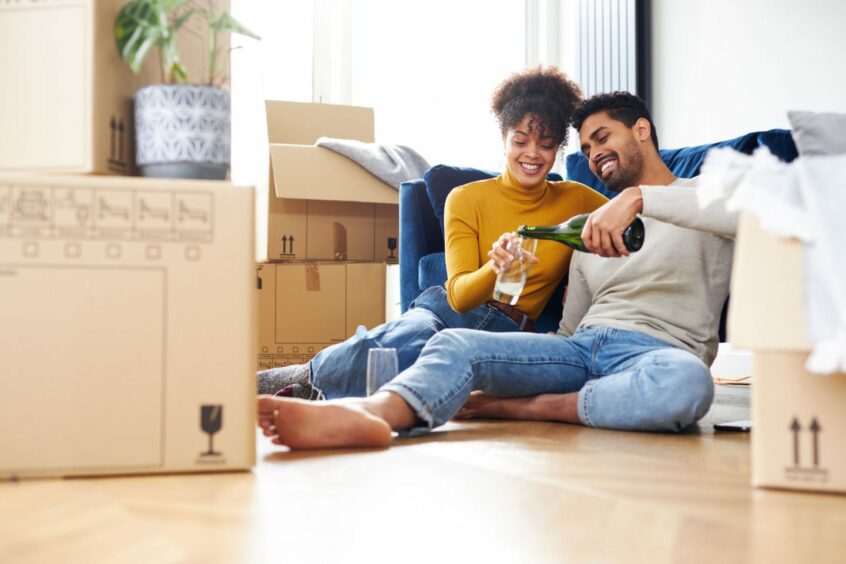 A couple sit on the floor and open a bottle of champagne to celebrate their house move while surrounded by boxes. 