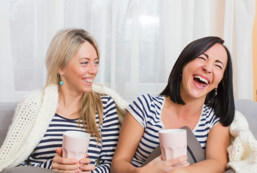 Two women sitting with blanket over their backs laughing while holding their cups of tea