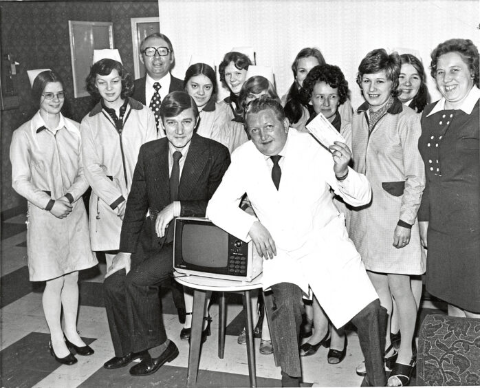 A group of staff members huddled around a portable television, with a man in a white coat holding up a cheque