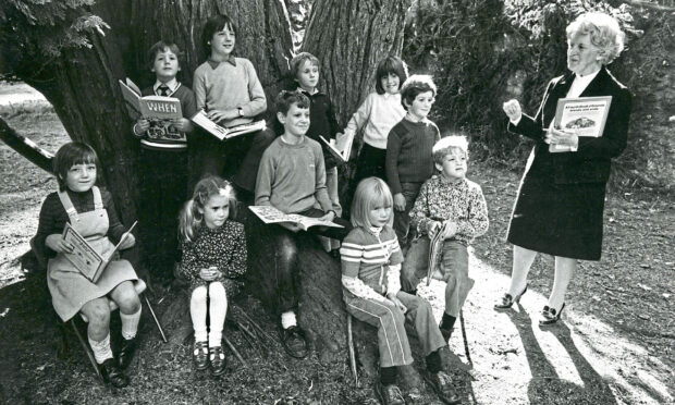 A teacher standing outside with a class of children sitting at the foot of a tree