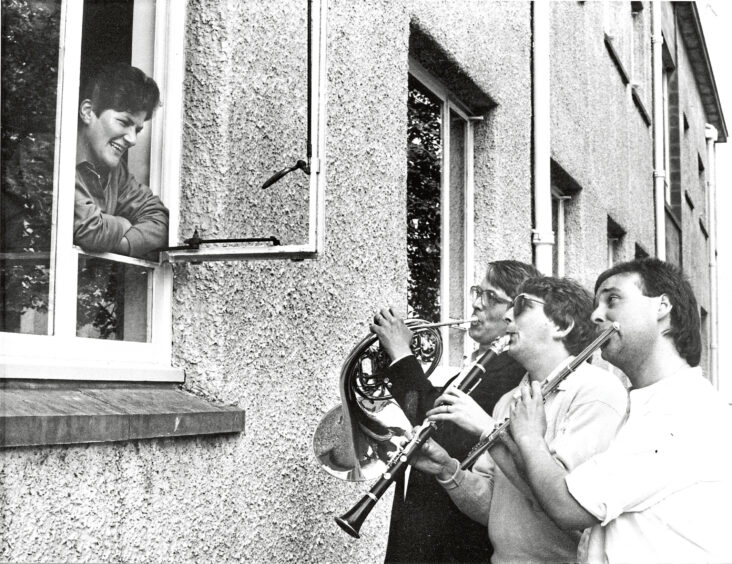 Three men play their instruments up to a man leaning out of a window