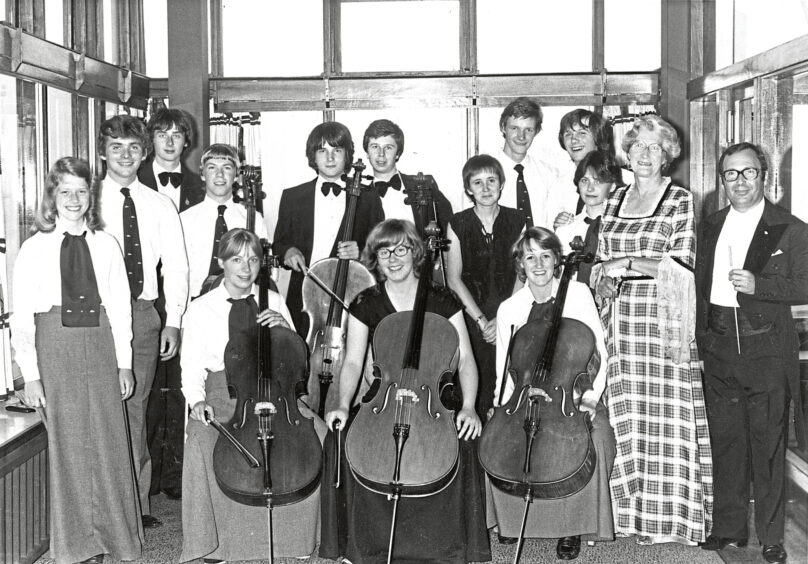 A large group smile at the camera, gathered around cellists at Aberdeen International Youth Festival