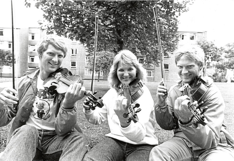 Three siblings hold their violins up to their chins and smile at the camera before performing at the Aberdeen International Youth Festival