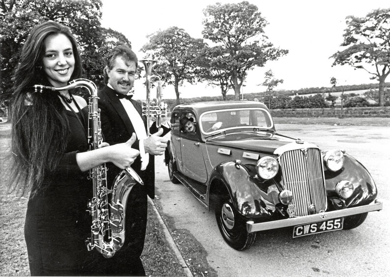 A man holding a trumpet and woman holding a saxophone put their thumbs up and smile at the camera with a car stopping beside them