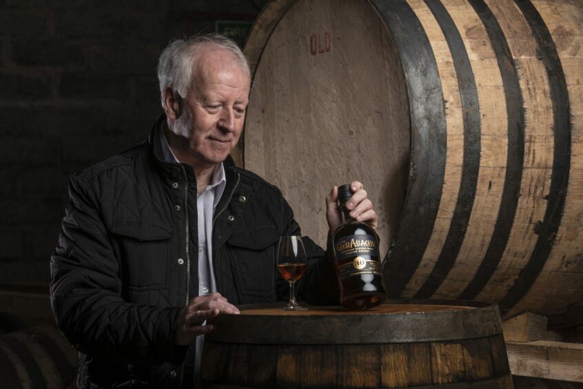 The GlenAllachie master distiller Billy Walker holding a bottle in front of whisky casks.
