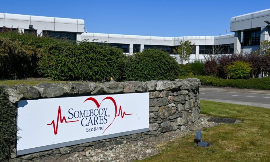 Sign on a brick wall that says "Somebody Cares Scotland". Bushes, trees and a building is in the background.