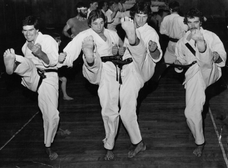 Four men in karate uniform in a line, kicking towards the camera