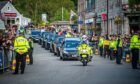 Looking head-on at Queen's coffin cortege headed by police motorbike in Ballater with packed streets either side.
