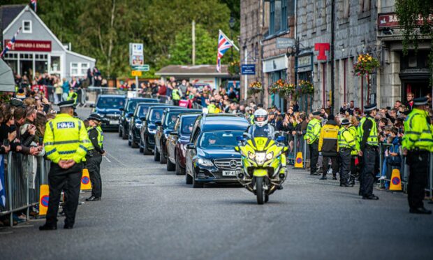 Looking head-on at Queen's coffin cortege headed by police motorbike in Ballater with packed streets either side.