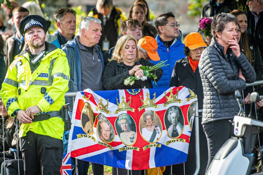 Crowd's wait patiently for the cortege in Ballater. A Union Jack with photographs of Queen Elizabeth II printed on it hangs from the barrier in front of them