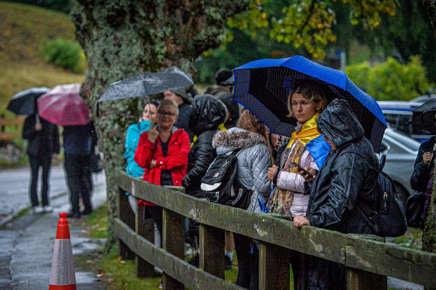 Group of people under umbrellas next to wooden fence next to side of road outside Balmoral
