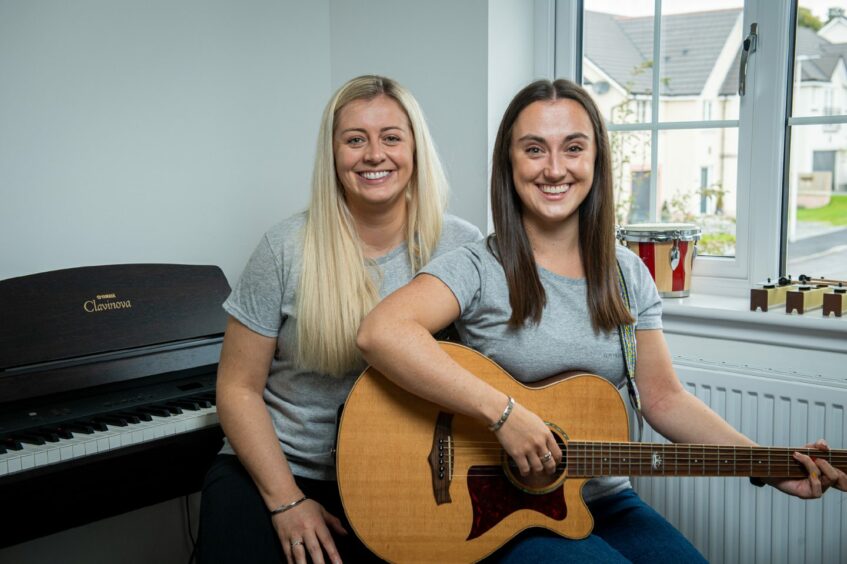 Emmeline McCracken sitting on stool next to piano and on the other stool Nadine Allan is holding her guitar as they smile for the camera