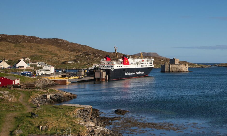 A fine day on Barra with a CalMac ferry and the castle in the bay at Castlebay on Barra, A few houses are also on the quayside with green rough rocks in the foreground. 