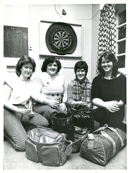 Four women sit in front of a dart board, smiling at the camera