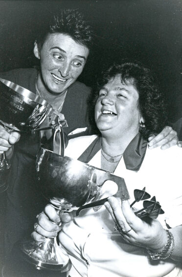 Two women embrace, smiling, while holding trophies and darts.